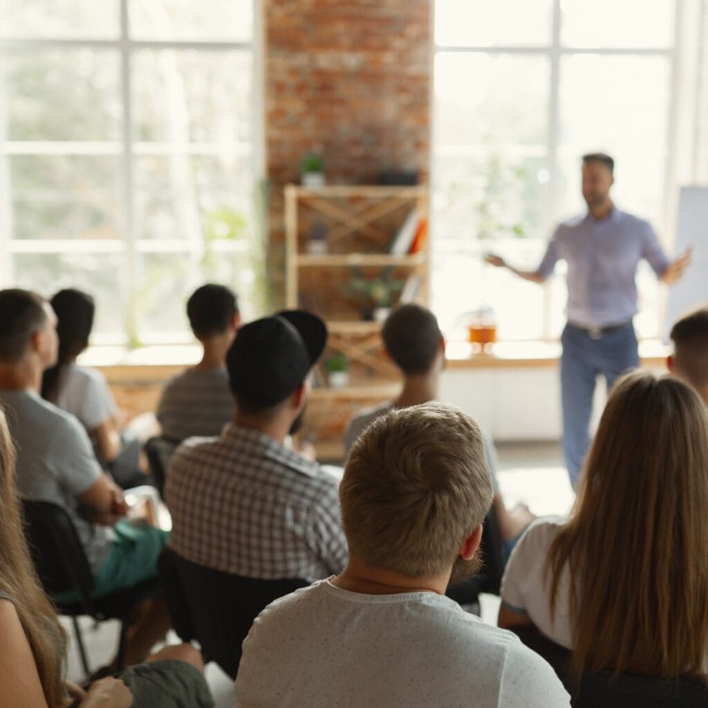 Male speaker giving presentation in hall at university workshop. Audience or conference hall. Rear view of unrecognized participants in audience. Scientific conference event, training. Education